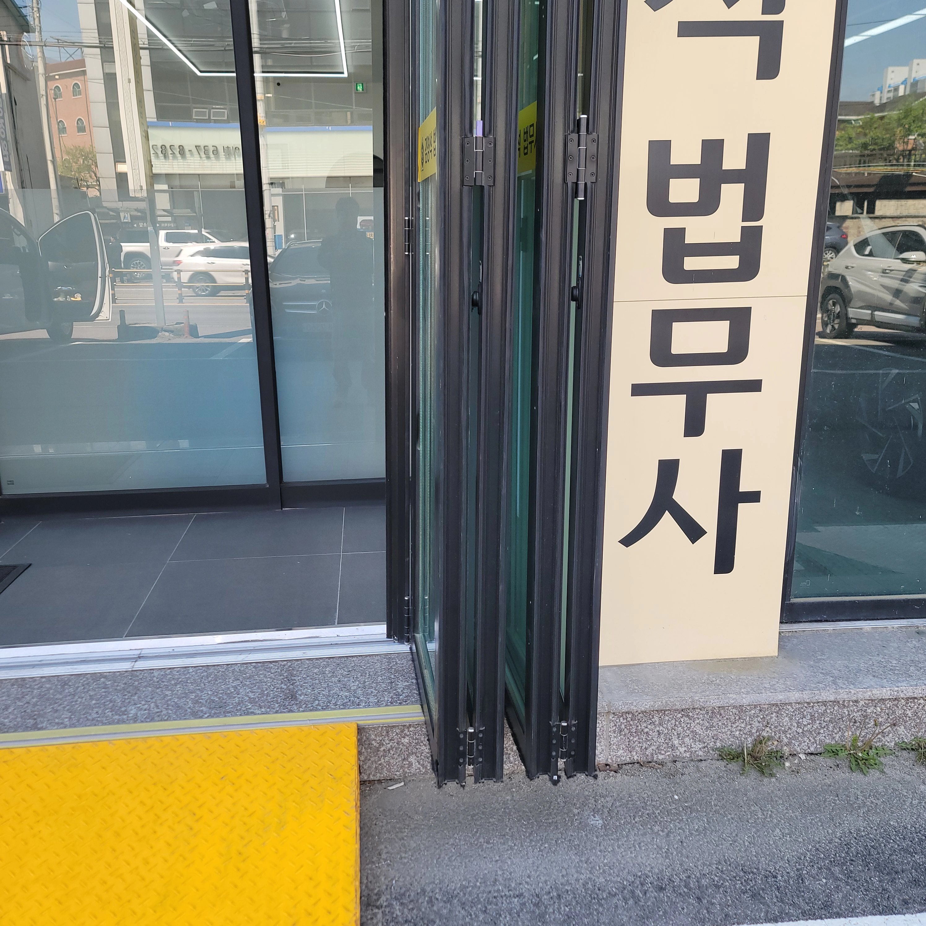 상가 건물 출입구에 설치된 접이식 유리도어로, 여러 짝의 유리 패널이 알루미늄 프레임에 경첩으로 연결되어 한쪽으로 접혀 열린 상태입니다.