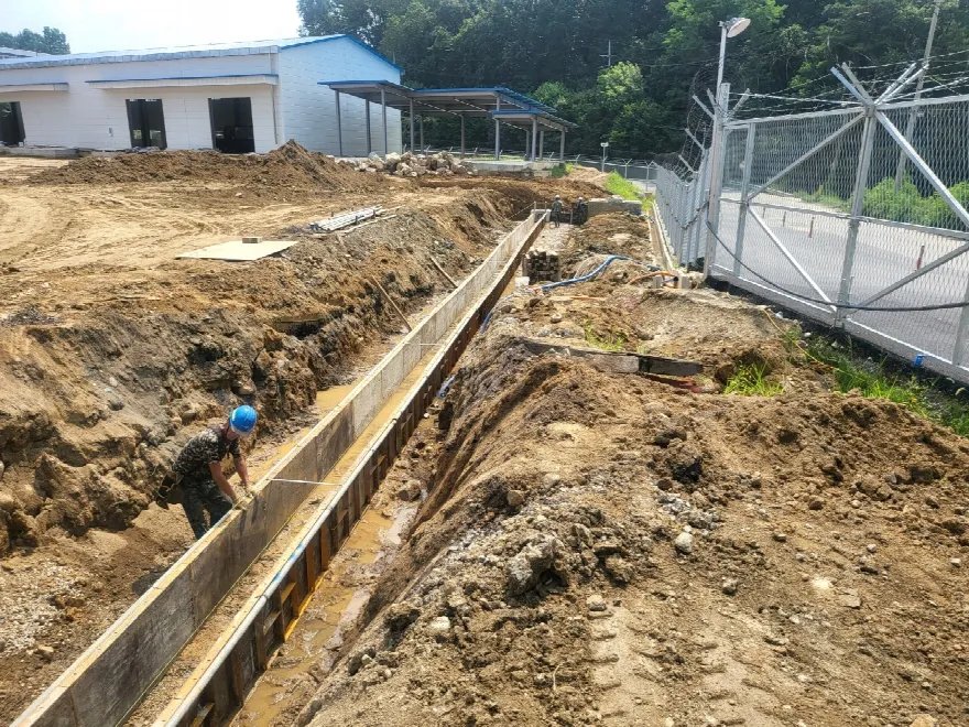 건물 뒤로 깊고 길게 판 도랑 안에 거푸집 판이 길게 세워져 있고, 작업자가 도랑 안에서 위치를 잡고 있습니다.