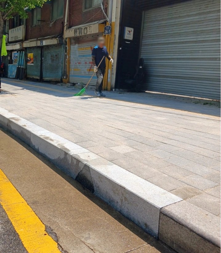 상가 앞 보도가 화강석 블록으로 새로 포장되어 있고 작업자가 빗자루로 바닥의 먼지와 부스러기를 쓸어내고 있다.