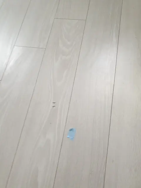 밝은 그레이 톤 강마루 한가운데 파란 보호 스티커가 임시로 붙어 있고 옆에 작은 점 자국 두 개가 보이는 시공 후 점검 사진