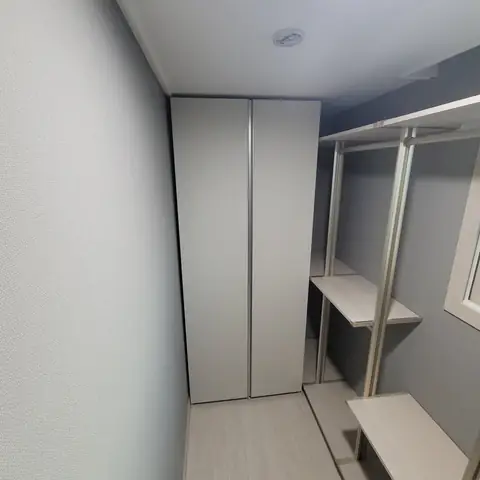좁은 드레스룸 안쪽에 회색 여닫이 붙박이장이 천장까지 서 있고 오른쪽 벽에 흰 기둥 프레임 선반과 옷걸이 봉이 달린 모습