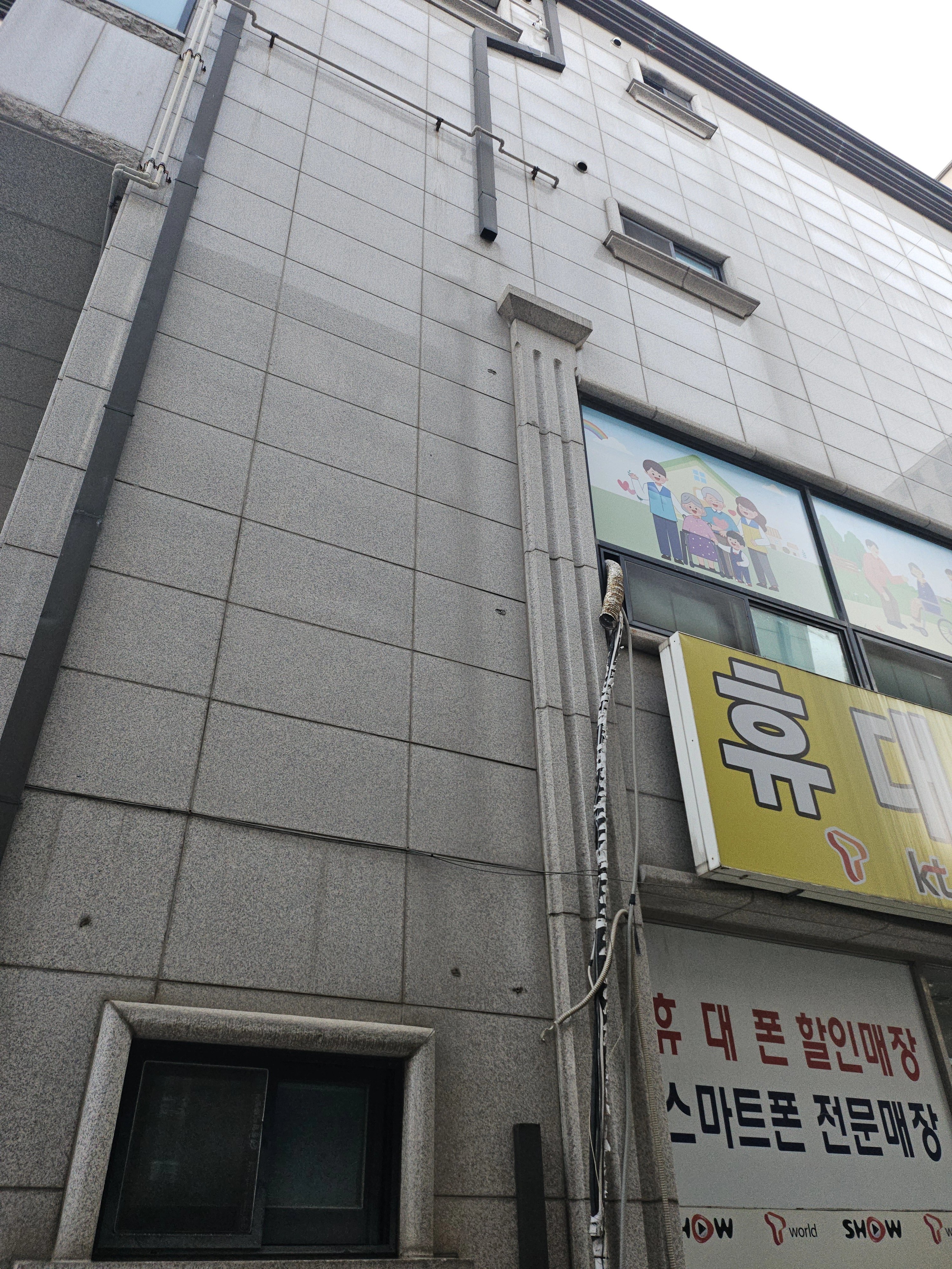 건물 외벽 타일 마감 사이로 흰색 우수관(추정)이 위에서 아래로 길게 내려와 있습니다.