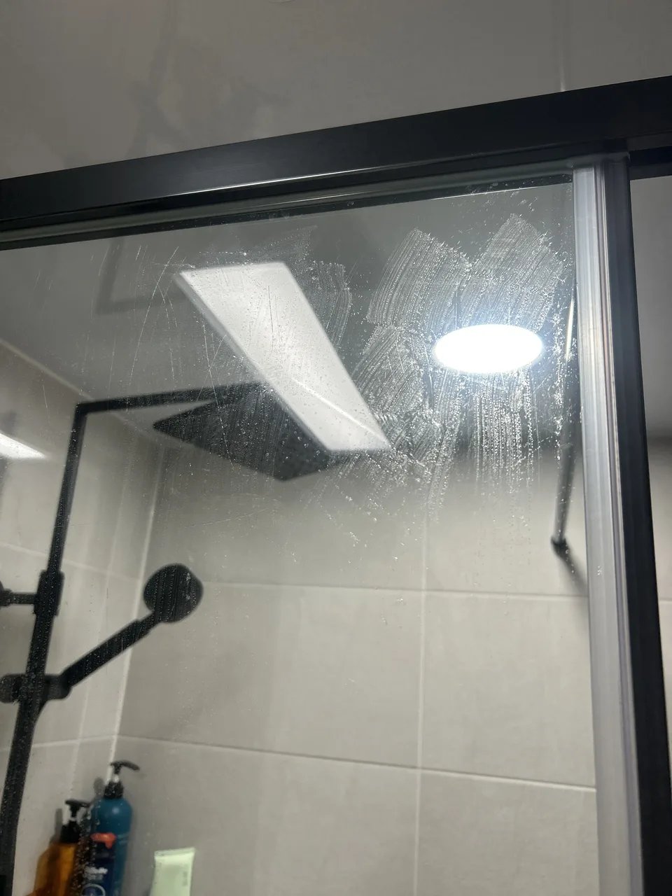 샤워부스 상단부를 가까이서 찍은 사진으로, 블랙 프레임 유리문 너머로 레인샤워 헤드가 설치된 모습이 보인다.