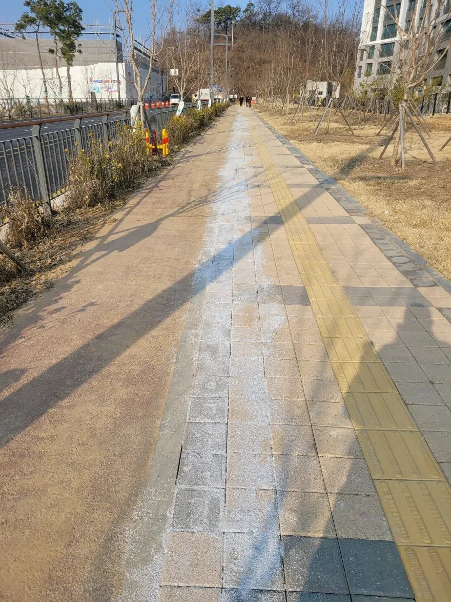 가로수와 펜스를 따라 길게 이어진 인도로, 회색 블록 구간과 노란 점자블록, 적갈색 포장이 나란히 놓여 있습니다.
