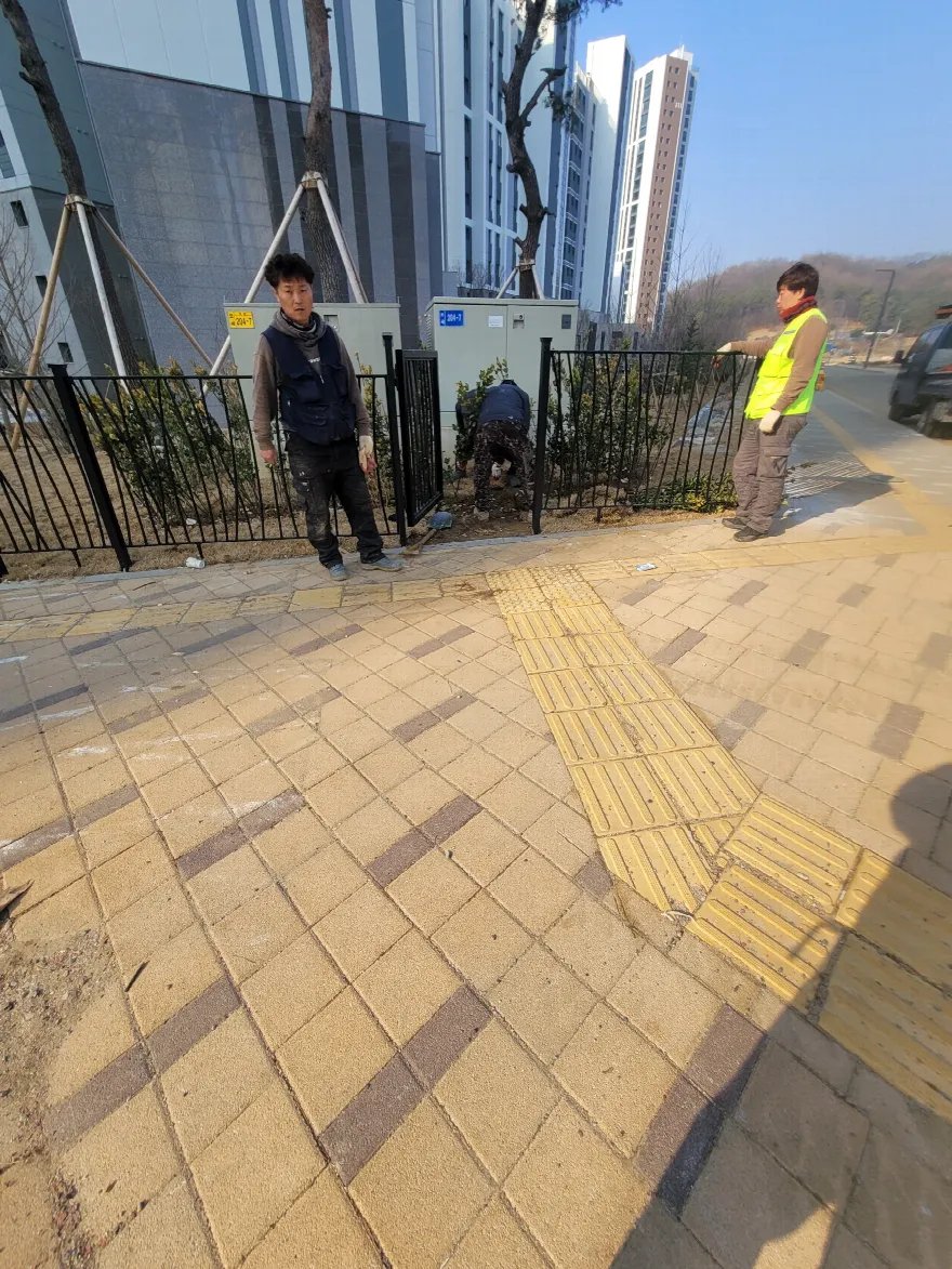 펜스와 화단 옆 인도에서 작업자들이 시공 구간을 살펴보는 모습입니다.