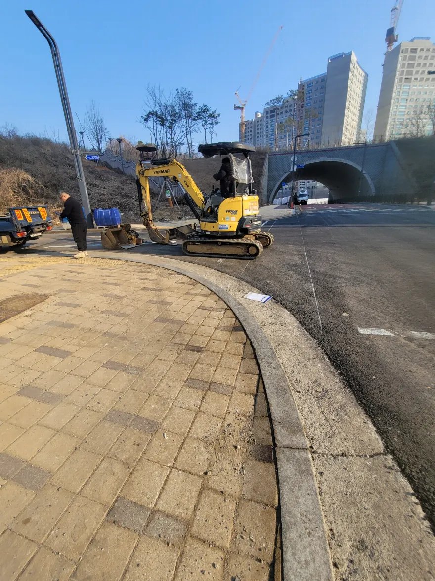 차도와 맞닿은 곡선 보도 모서리 구간에 소형 굴삭기가 진입해 작업을 준비하는 장면입니다.