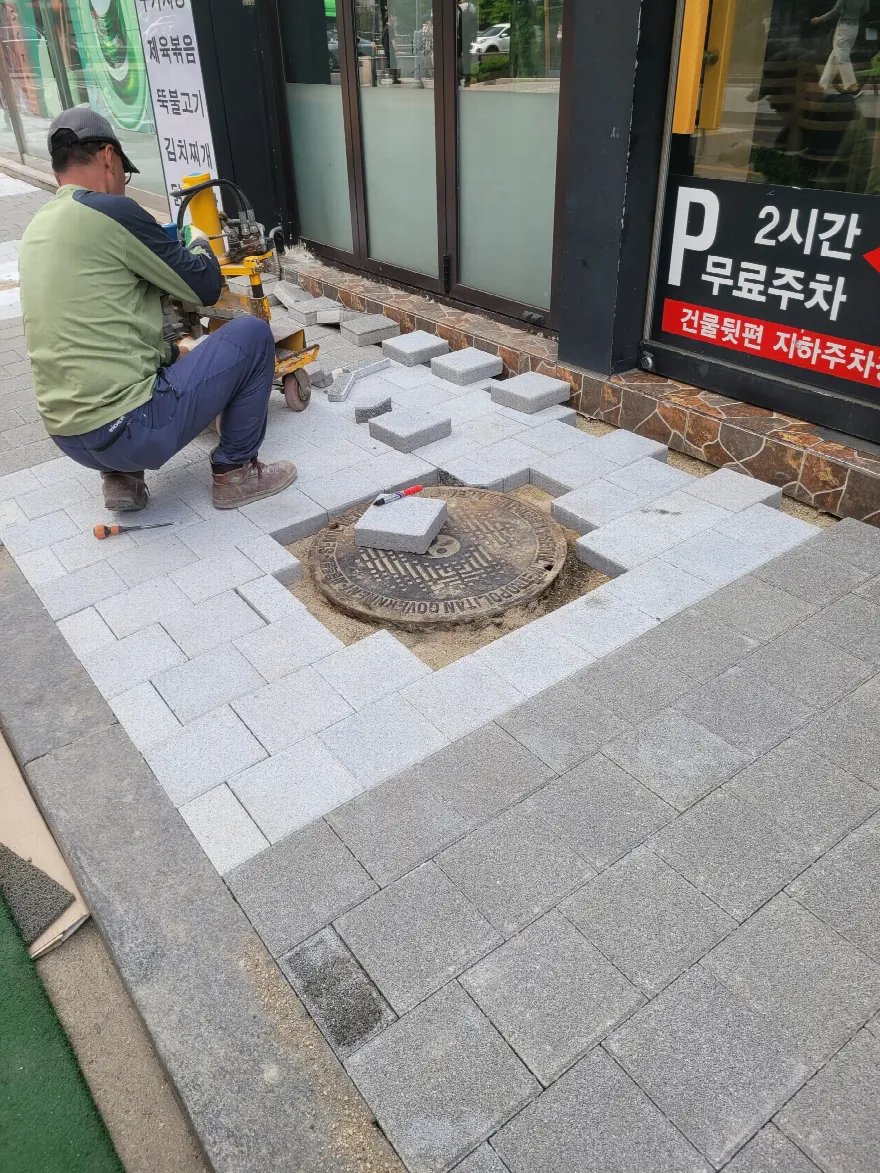 상가 앞 인도에 회색 화강 블록을 깔아 나가는 중이며, 원형 맨홀 뚜껑 주변을 남겨 두고 블록을 채워 가고 있습니다.