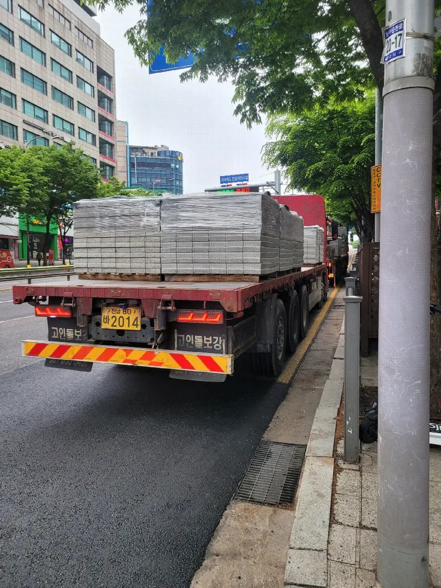 대형 화물차 적재함에 회색 보도블록이 팔레트 단위로 여러 단 실려 도착한 모습입니다.