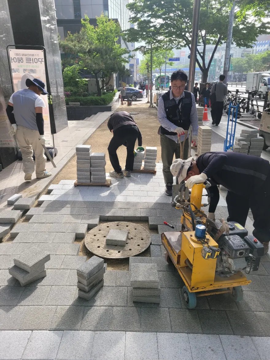 건물 앞 인도의 블록 깔기가 상당 부분 진행된 모습으로, 앞쪽 원형 맨홀 주변만 남겨 두고 회색 블록이 채워져 있습니다.