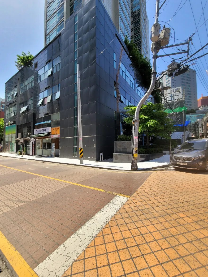 건물 모퉁이 앞 교차로에 붉은 벽돌색 블록 포장과 노란 유도 띠가 깔린 모습이 보입니다.
