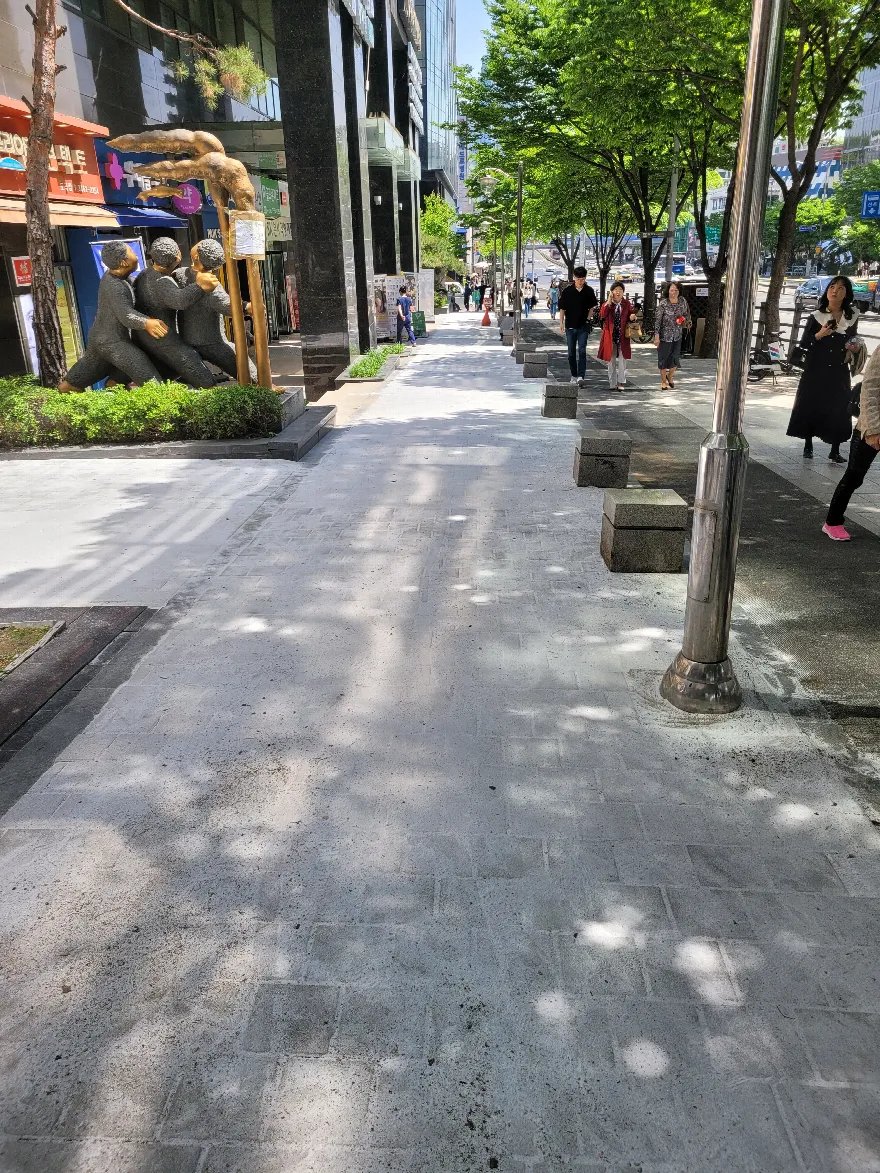 가로수가 늘어선 도심 보도에 큰 규격의 밝은 판석 블록이 깔려 있는 현장입니다.