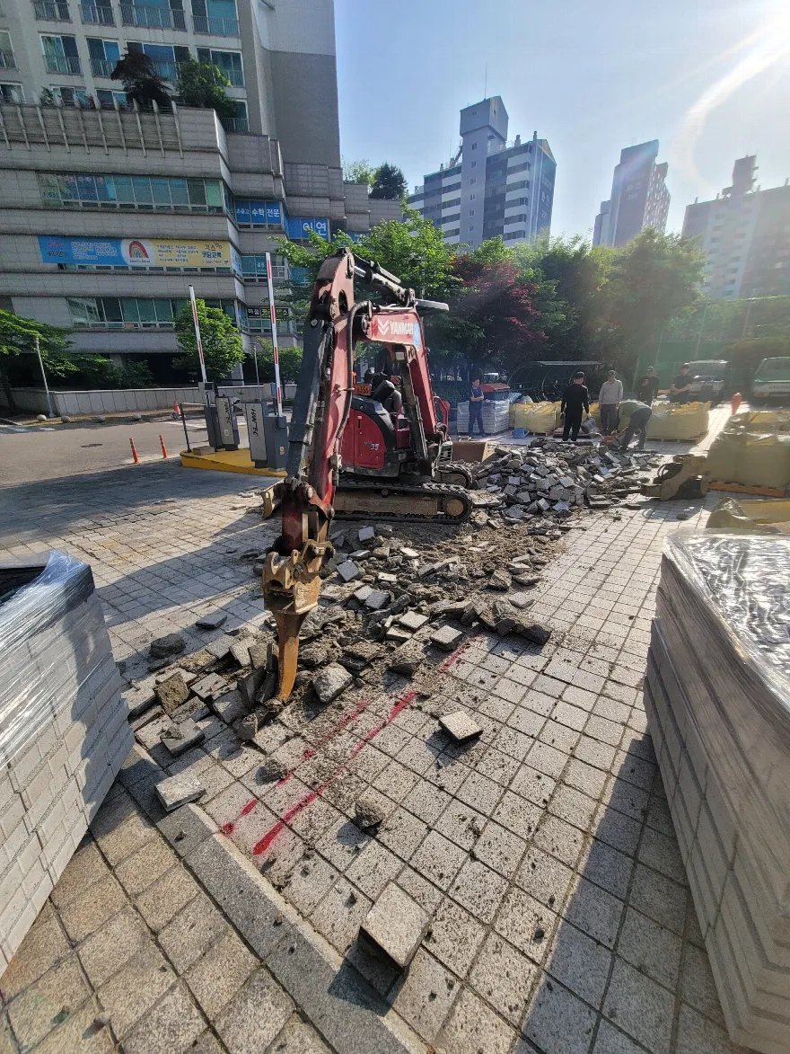 건물 앞 보도에서 소형 굴삭기로 기존 판석 블록을 걷어내는 중입니다.