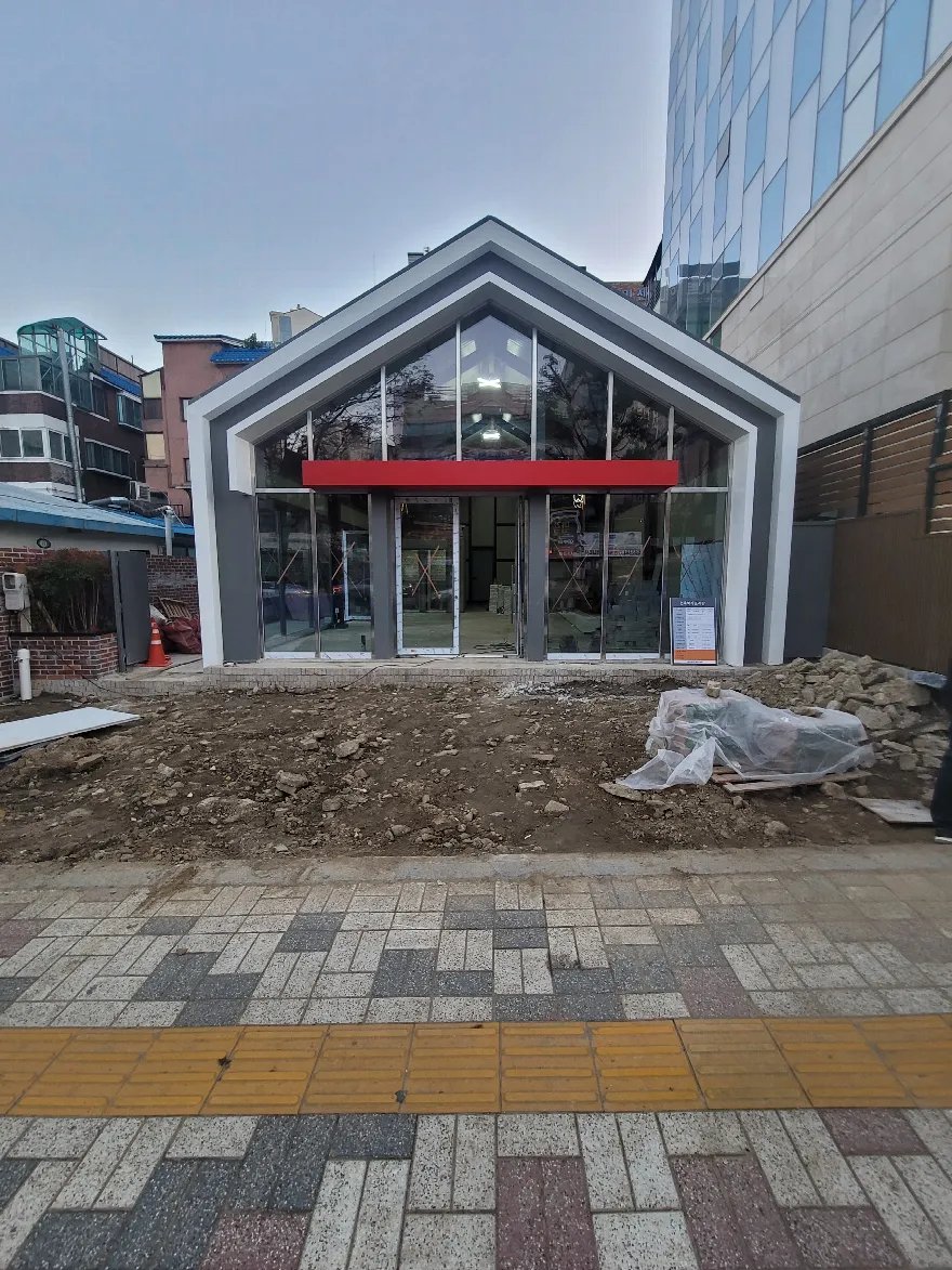 박공 지붕 건물 앞마당의 기존 바닥을 걷어내어 흙과 콘크리트 부스러기가 드러난 상태이며, 한쪽에는 비닐로 덮어 둔 자재가 있습니다.