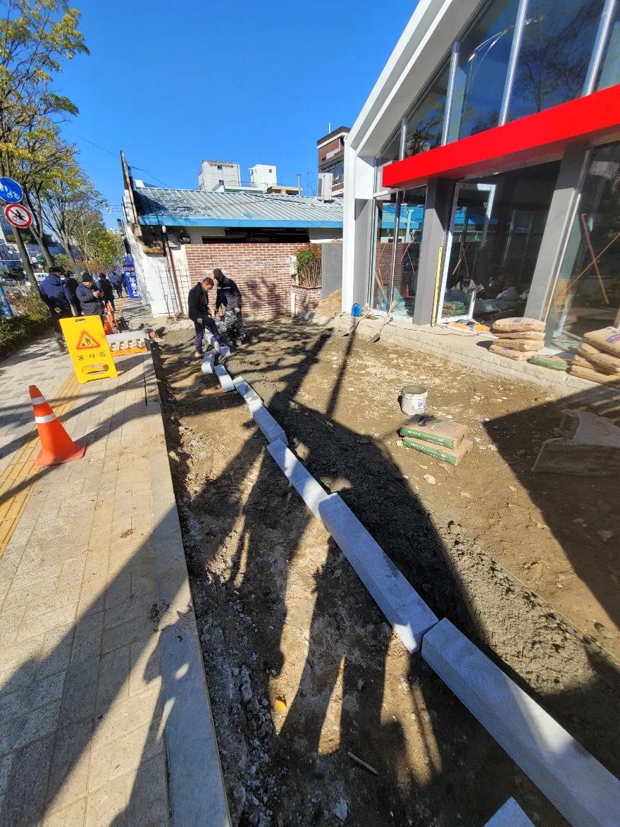 건물 앞 굴착한 바닥 가장자리에 흰 경계석을 줄 맞춰 세우고 있으며, 작업자들이 경계석을 내려놓고 위치를 잡는 중입니다.