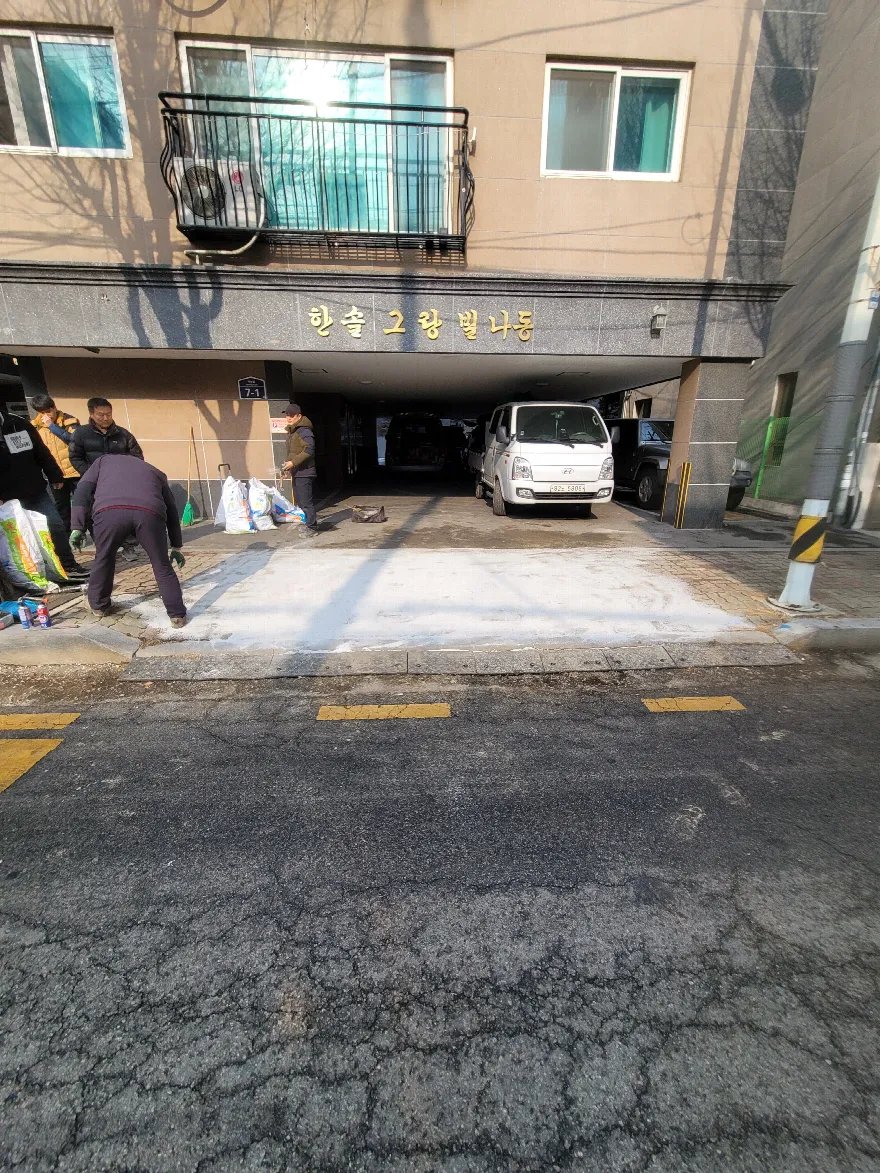 건물 필로티 주차장 진입로 앞 바닥에 밝은 색 모래 또는 몰탈층이 넓게 펴져 있고 작업자들이 가장자리에서 정리 작업을 하고 있습니다.