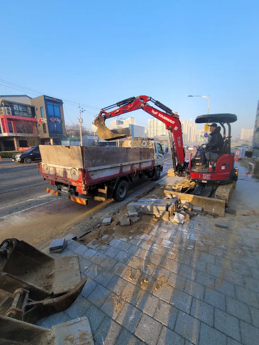 소형 굴삭기로 걷어낸 기존 보도블럭과 경계석 파편을 트럭에 상차하고 있는 철거 현장입니다.