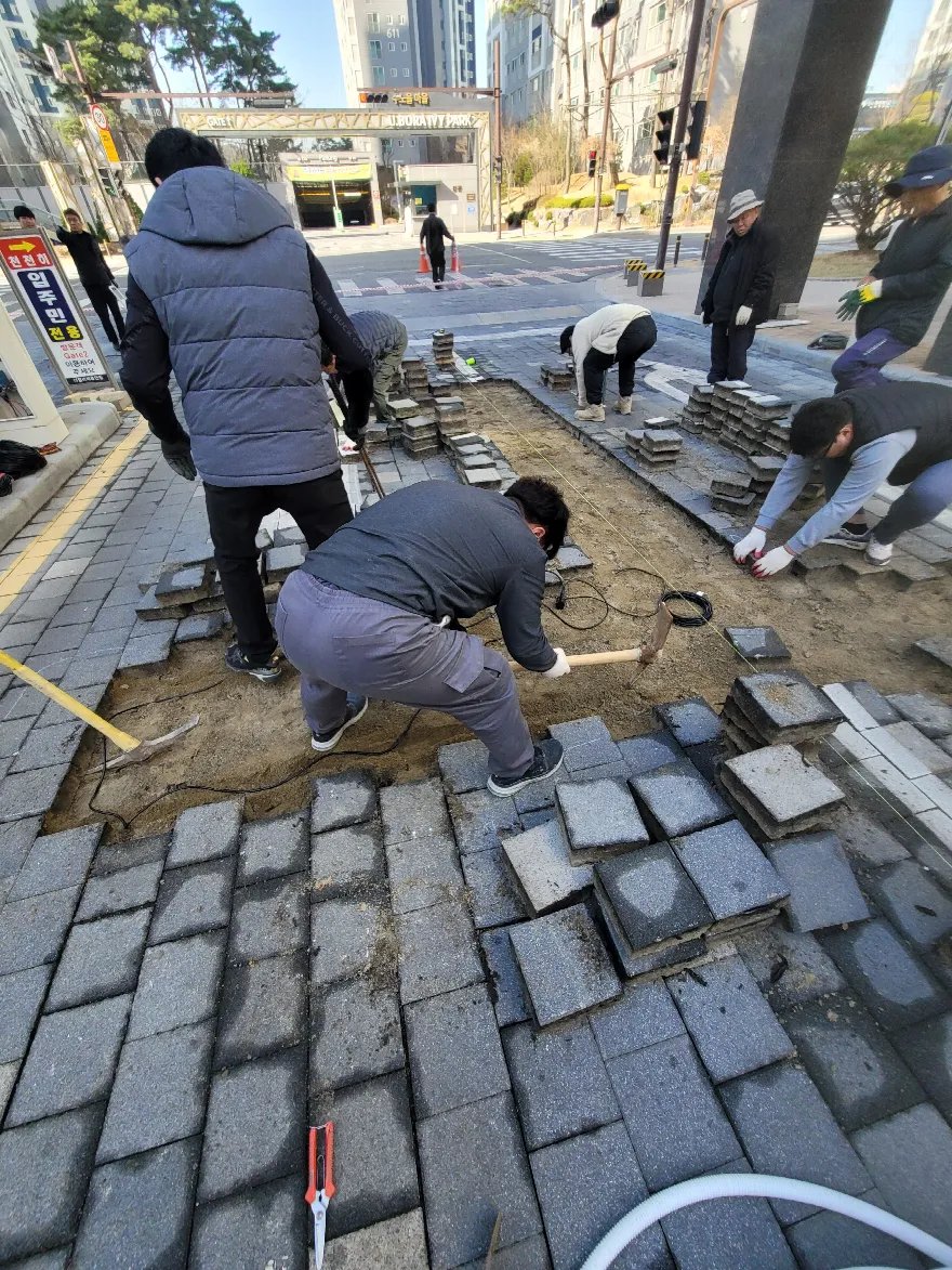 단지 출입구 보도에서 여러 작업자가 회색 사각 블럭을 다시 깔고 있는 장면입니다.