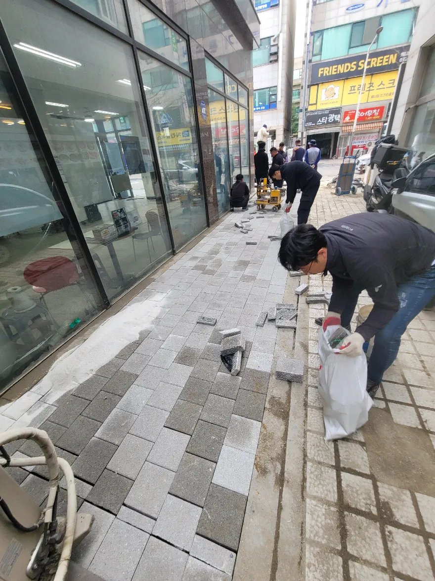 상가 유리 외벽 앞 보도에 밝은 회색과 진회색 사각 블록을 섞어 깔고 있는 중입니다.