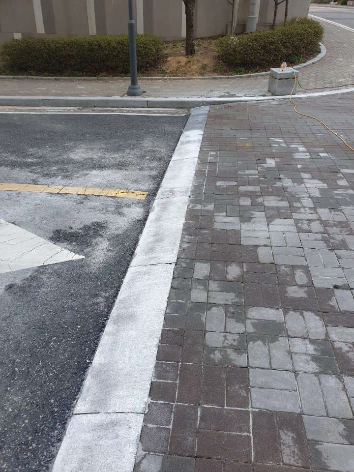 아스팔트 차도와 맞닿은 보도에 진회색 블록 포장이 되어 있고, 경계석은 흰색으로 마감되어 있습니다.