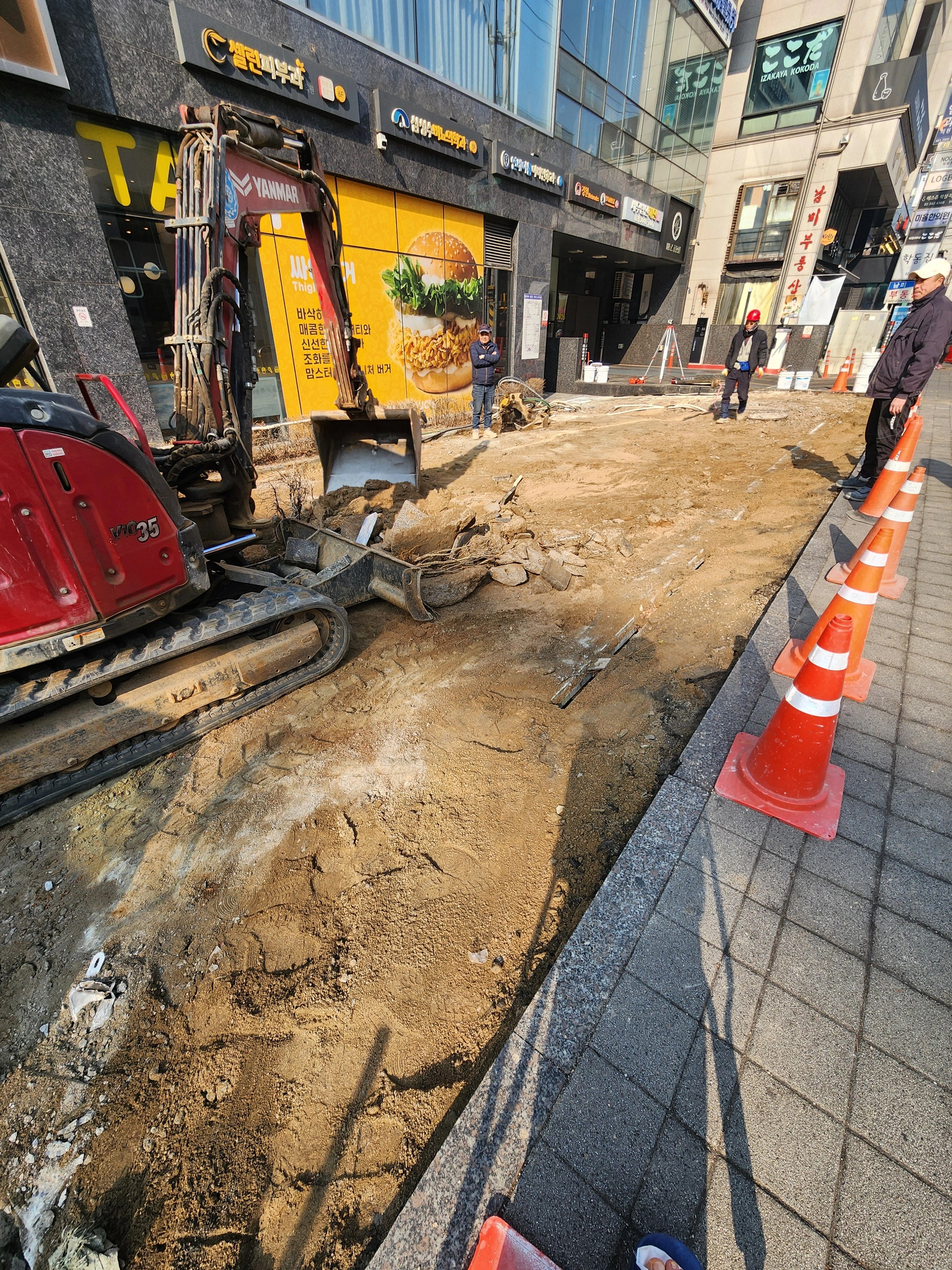 상가 건물 앞 보도 구간의 기존 블록을 걷어내고 미니굴착기로 하부 바닥을 정리하는 작업이 진행 중입니다.