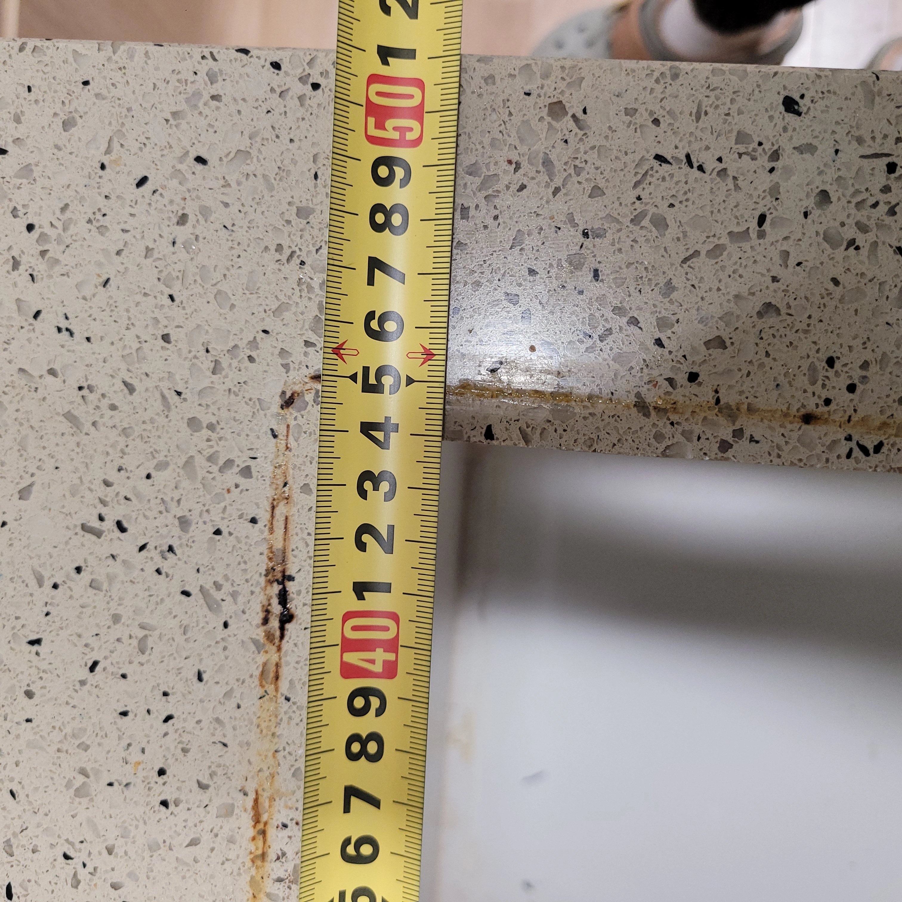 돌가루가 섞인 인조대리석(또는 테라조) 상판 모서리에 줄자를 대고 치수를 재는 장면입니다.