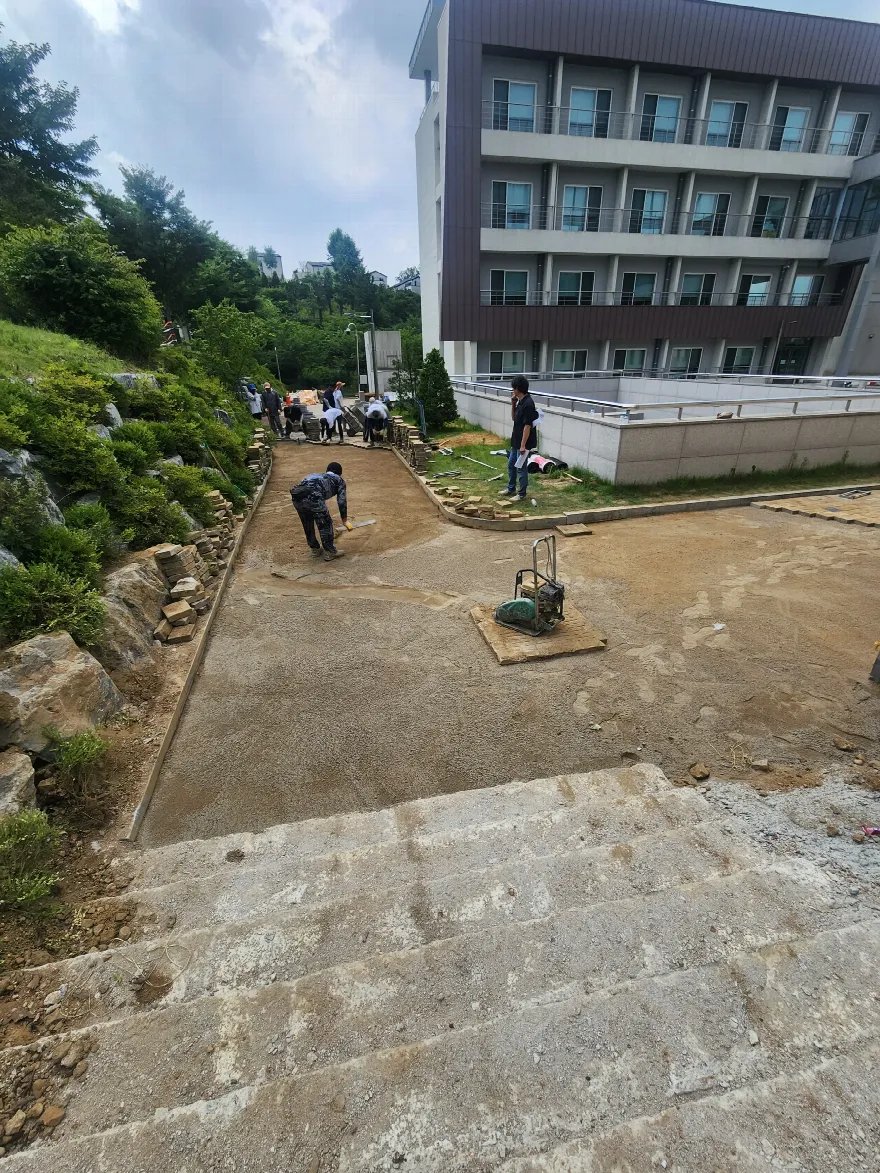 건물 앞 경사 통로의 흙 바탕을 작업자들이 갈퀴와 삽으로 고르고 있고, 가운데에 다짐기가 놓여 있어 포설 후 다짐이 이어질 단계입니다.