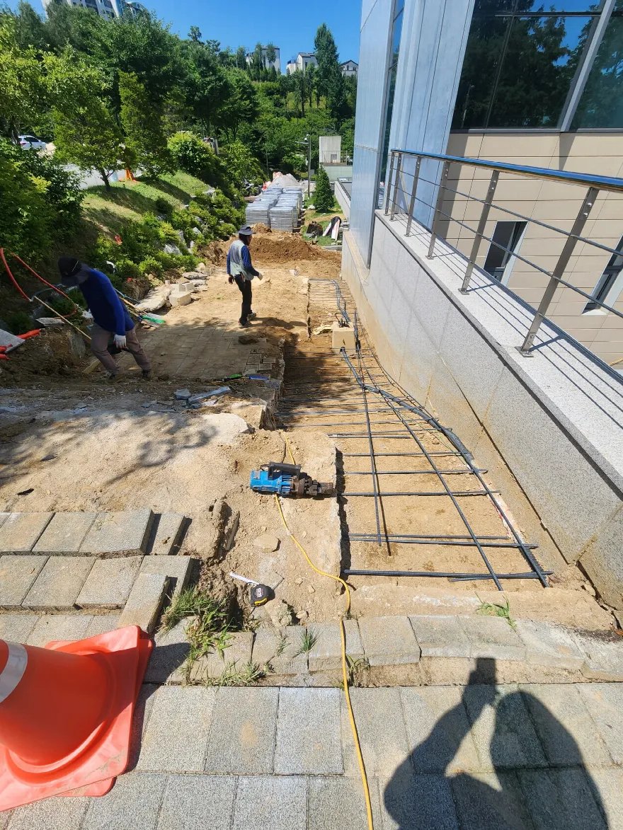 건물 외벽 옆 보행로 구간에 철근을 격자로 배근해 놓은 모습이고, 작업자 두 명이 안쪽에서 흙 정리를 하고 있습니다.