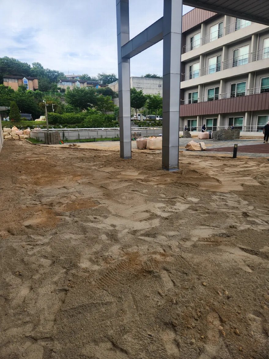 건물 앞 마당의 포장을 걷어내고 마사토 바닥이 넓게 드러난 상태로, 기둥이 서 있는 캐노피 아래까지 정지 작업이 되어 있습니다.