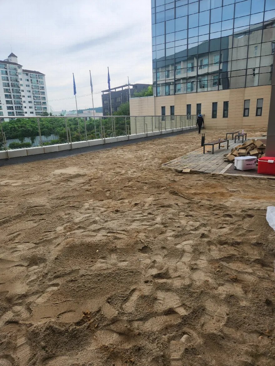 유리 난간으로 둘러싸인 옥상 테라스 바닥 전체에 모래를 포설한 준비 단계 사진입니다.