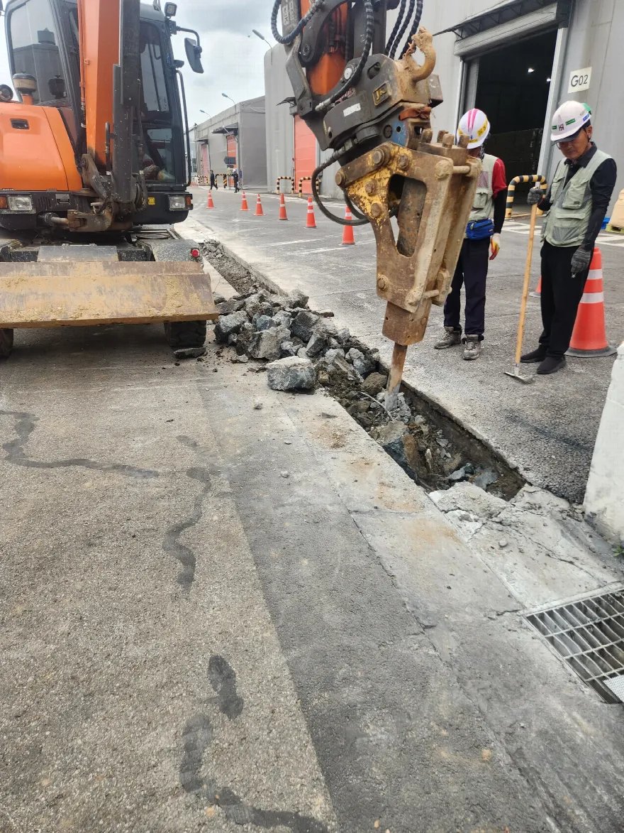 건물 출입구 앞 경계석을 따라 브레이커로 콘크리트 바닥을 깨고 있는 장면입니다.